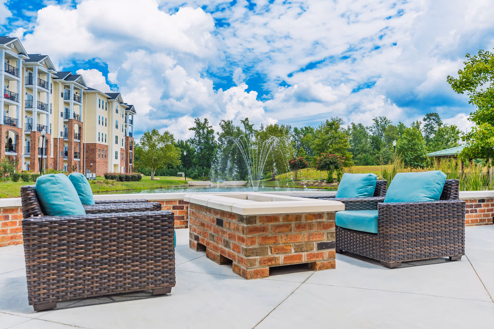 Outdoor seating area with four wicker chairs featuring turquoise cushions arranged around a brick fire pit. In the background, there is a pond with a water fountain, green trees, and a multi-story residential building under a partly cloudy blue sky.