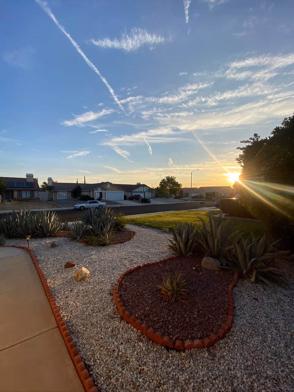 A landscaped front yard with decorative gravel and plants bordered by red bricks, a sidewalk on the left, a street with houses in the background, and the sun setting near the horizon with a partly cloudy sky.