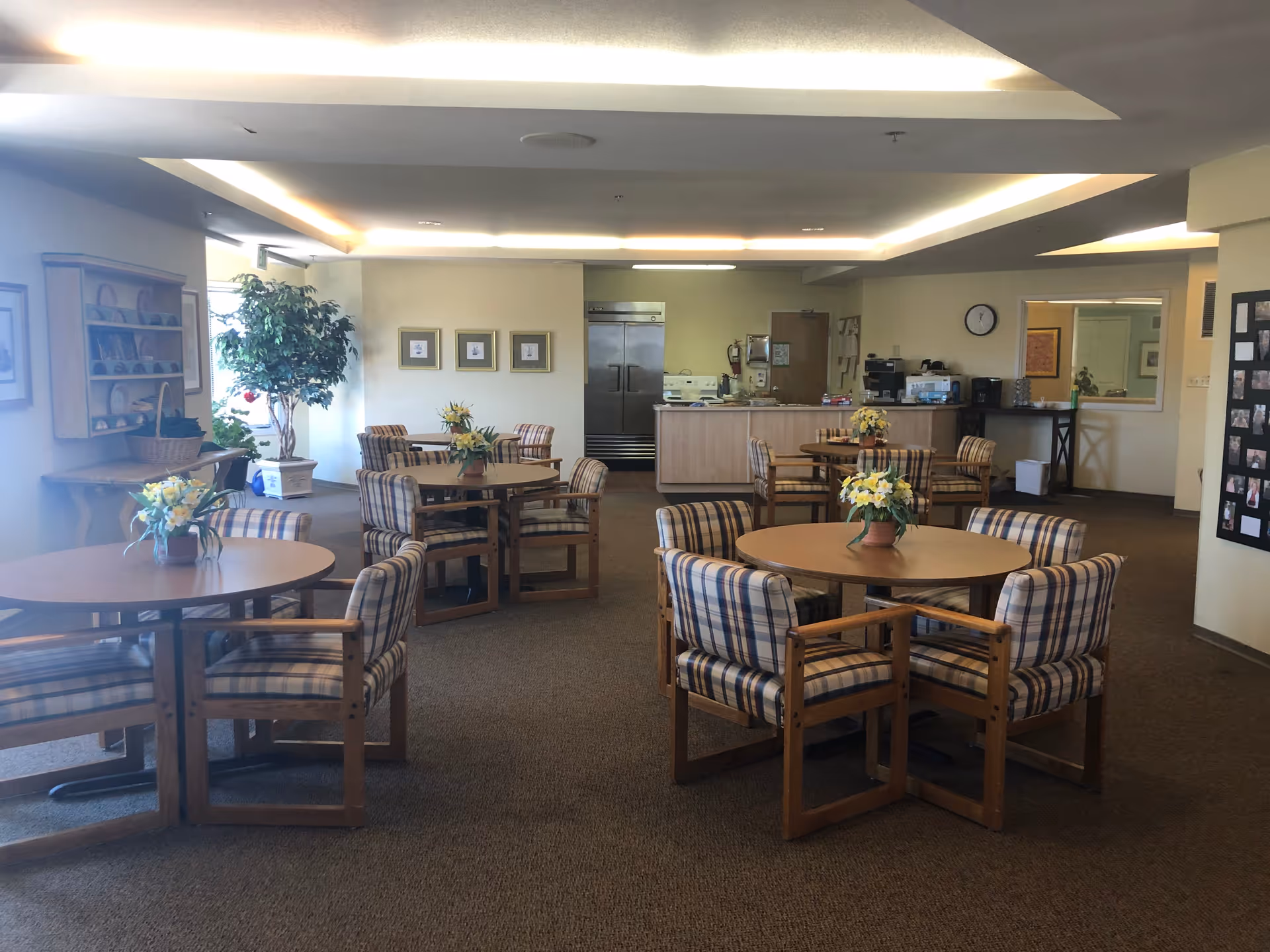 A senior living facility common dining area with round wooden tables and plaid cushioned chairs. Each table has a flower arrangement. The room has beige walls, carpeted floor, and a ceiling with recessed lighting. In the background, there is a kitchenette area with a refrigerator, stove, and coffee machine. A large potted plant and framed pictures are visible on the walls.