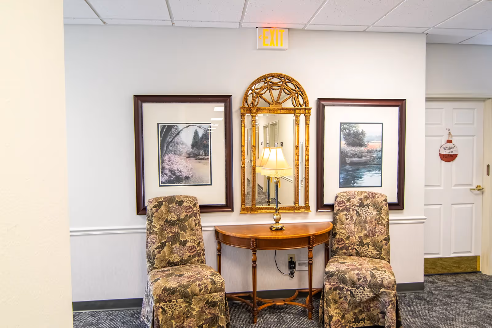 A hallway seating area with two floral upholstered chairs on either side of a wooden console table. On the table is a lamp with a beige shade. Above the table is an ornate gold-framed mirror flanked by two framed landscape paintings. A white door with a hanging sign that reads 'Wishin' I was Fishin'' is visible to the right. An illuminated exit sign is mounted on the wall above the mirror.