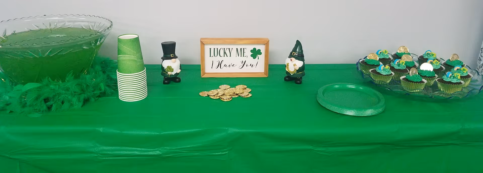 A green-themed party table with a large punch bowl filled with green drink, stacked green paper cups, two small leprechaun figurines, a sign that reads 'LUCKY ME, I Have You!', a pile of gold chocolate coins, a green plate, and a tray of decorated cupcakes with green frosting and gold coins on top.