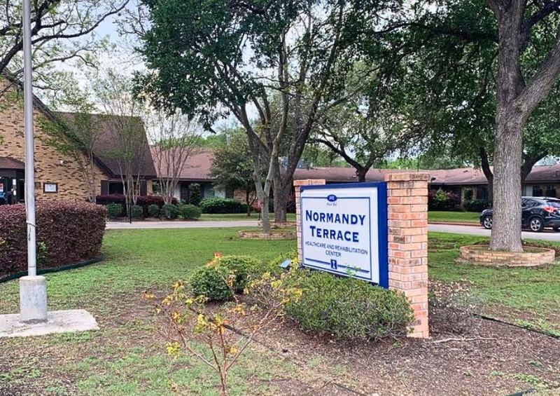Outdoor view of Normandy Terrace Healthcare and Rehabilitation Center showing a brick sign with the facility name surrounded by grass, bushes, and trees. The building and parking area are visible in the background.