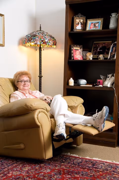 An elderly woman with glasses sits comfortably in a beige recliner chair with her legs extended on the footrest. She is in a cozy living room area with a colorful stained glass floor lamp beside her and a wooden bookshelf behind her displaying framed photos and decorative items.