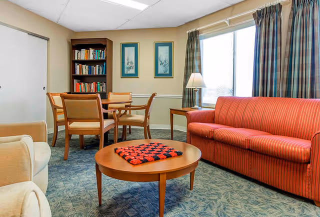 A cozy living room area with a red striped sofa, a round wooden coffee table with a red and black crocheted blanket on it, a beige armchair, a wooden dining table with four chairs, a bookshelf filled with books, two framed pictures on the wall, a side table with a lamp, and a large window with plaid curtains.