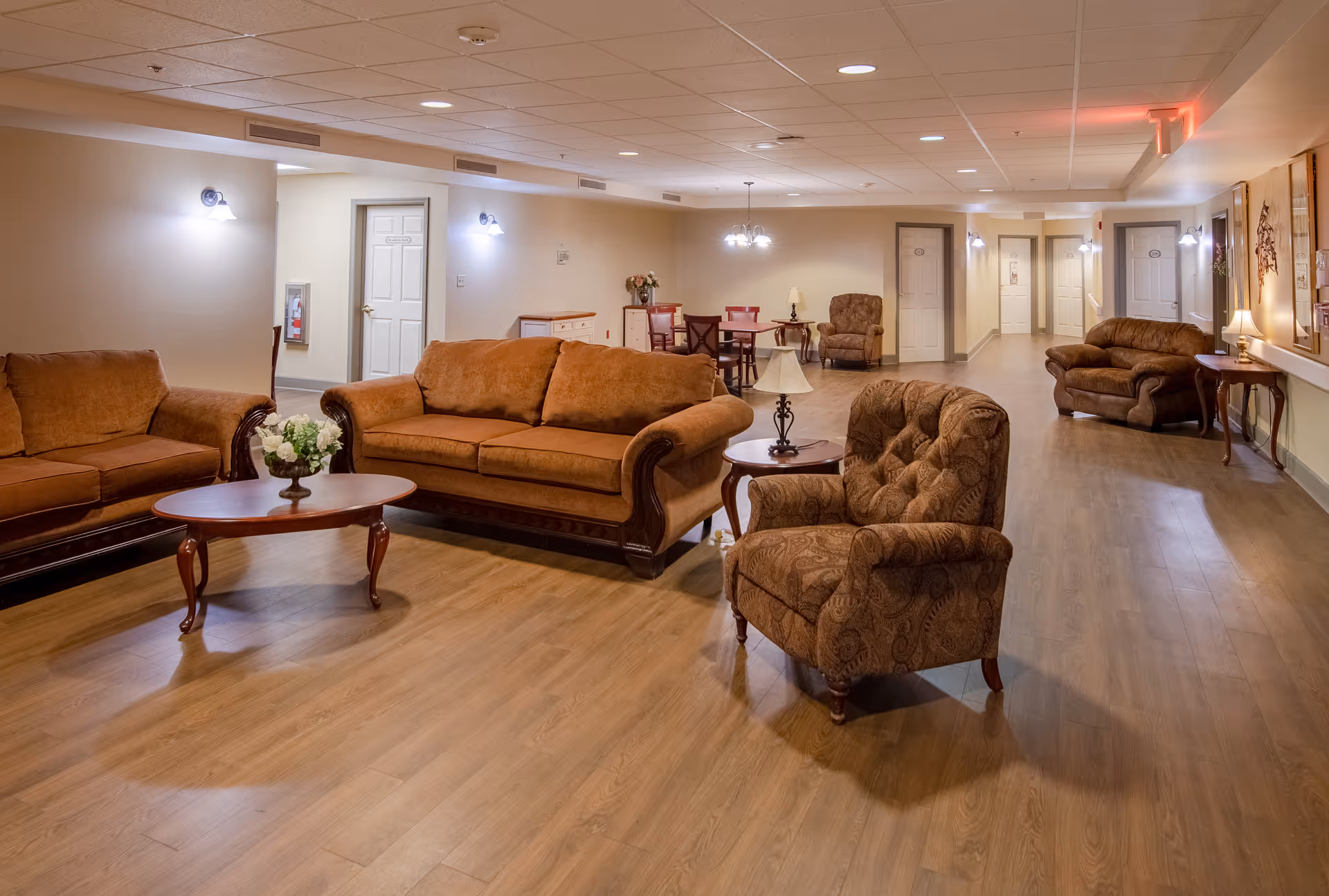A spacious, well-lit common area with brown upholstered sofas and armchairs arranged around wooden coffee tables. The room has wood flooring, beige walls, multiple doors, wall-mounted lights, and a small dining table with chairs in the background.