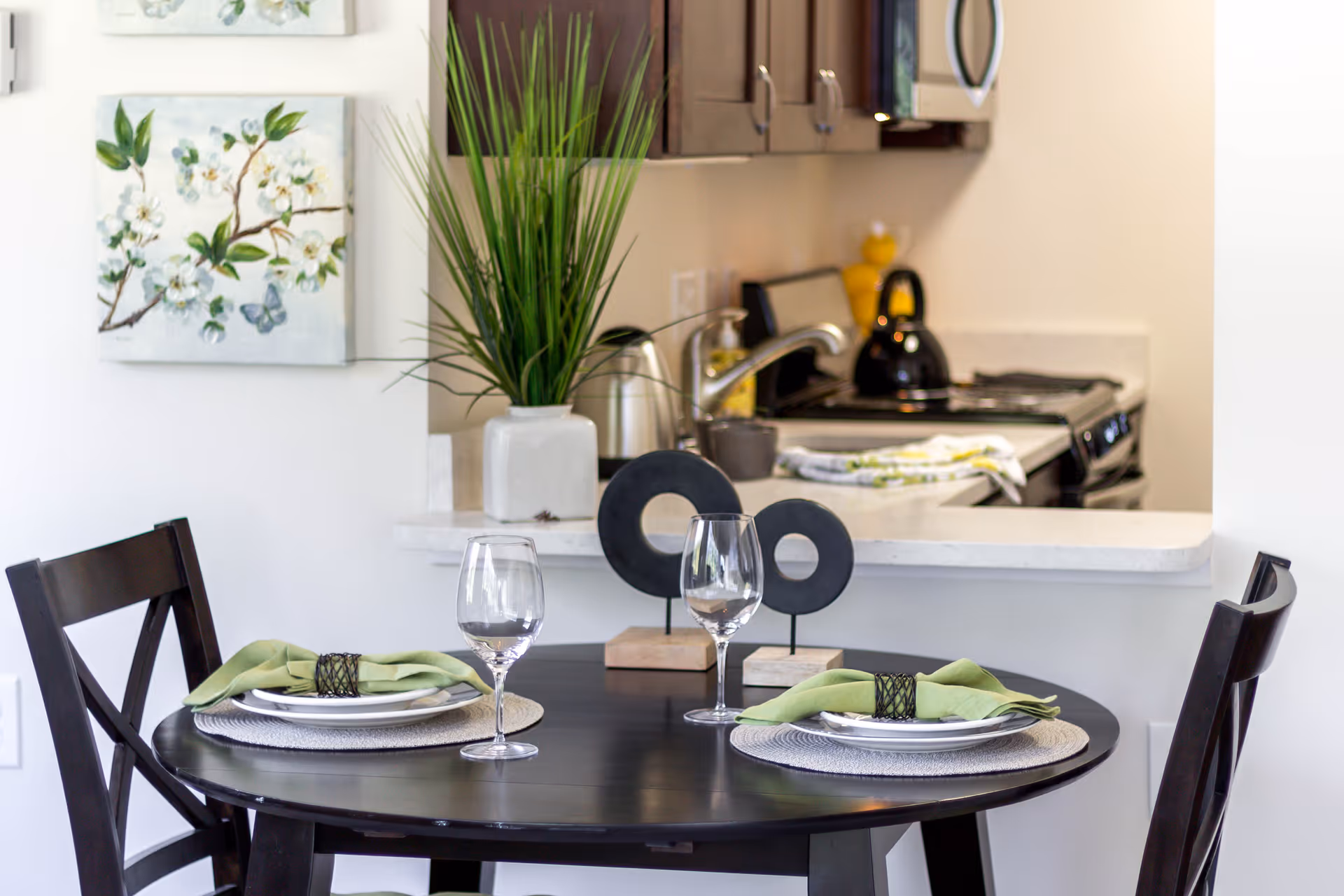 Round dining table set for two with wine glasses and green napkins in a dining area opening to a kitchen with dark cabinets and a potted plant.