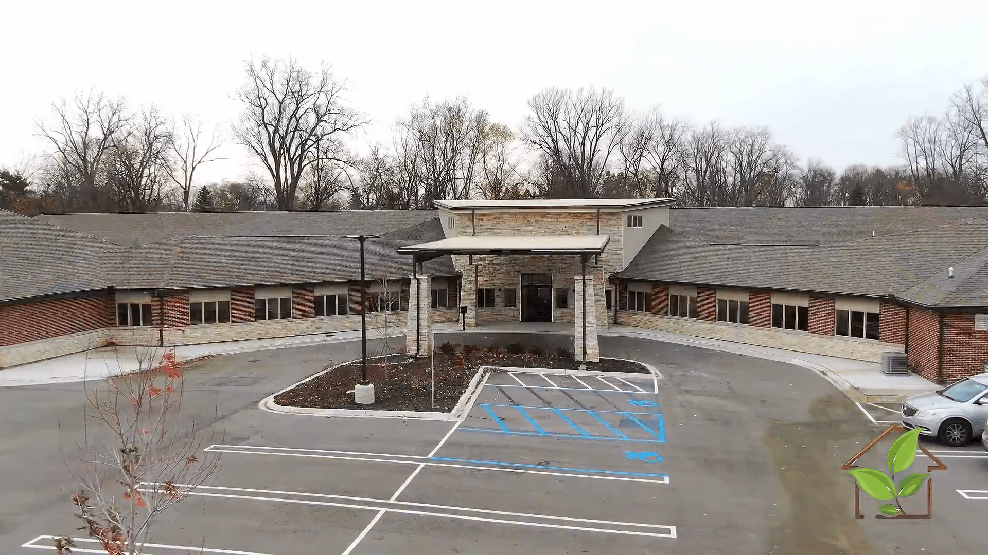 Exterior view of a single-story senior living facility building with a covered entrance, surrounded by a parking lot with marked parking spaces including handicapped spots. Leafless trees are visible in the background under an overcast sky.