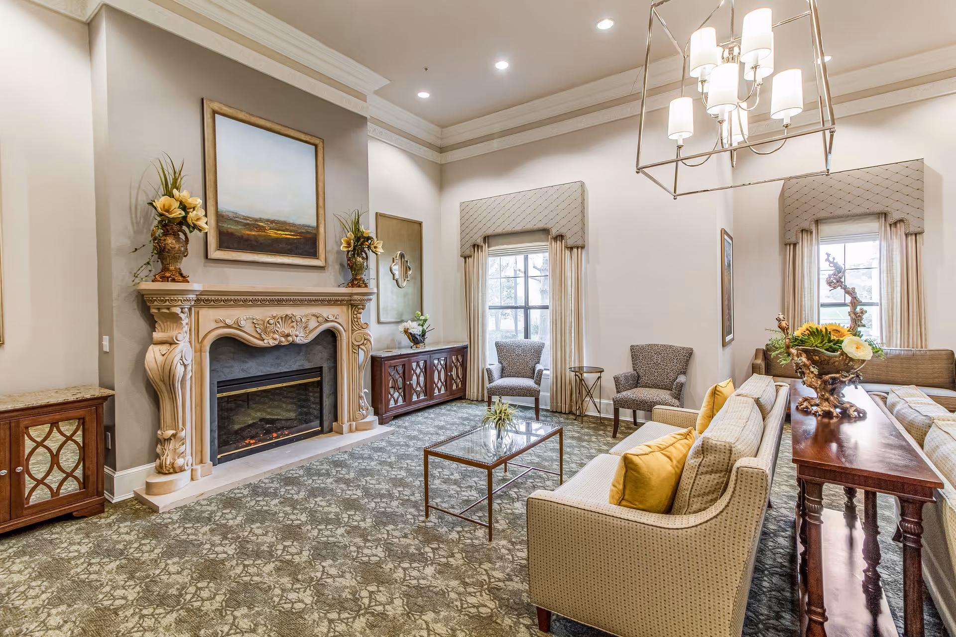 Spacious, well-lit communal living room with an ornate fireplace, sofas, armchairs, a glass coffee table and chandelier.