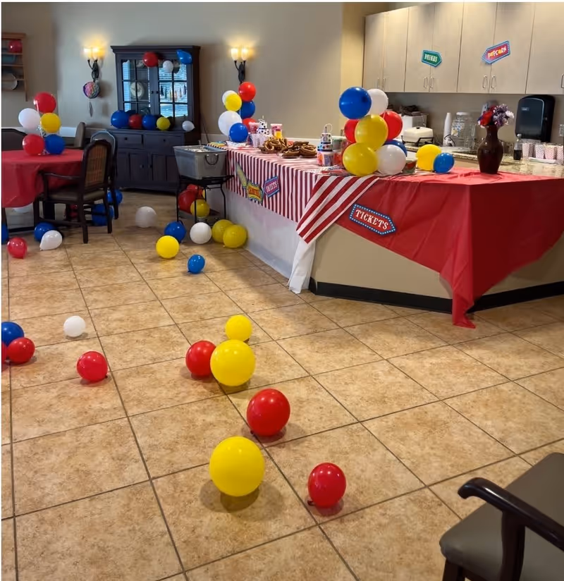 Interior room decorated for a celebration with colorful red, yellow, blue, and white balloons scattered on the tiled floor and arranged in clusters on tables. A long counter is covered with red and white striped tablecloths and has snacks, pretzels, and cups on it. The room has beige walls, a dark wooden cabinet, and chairs around tables.