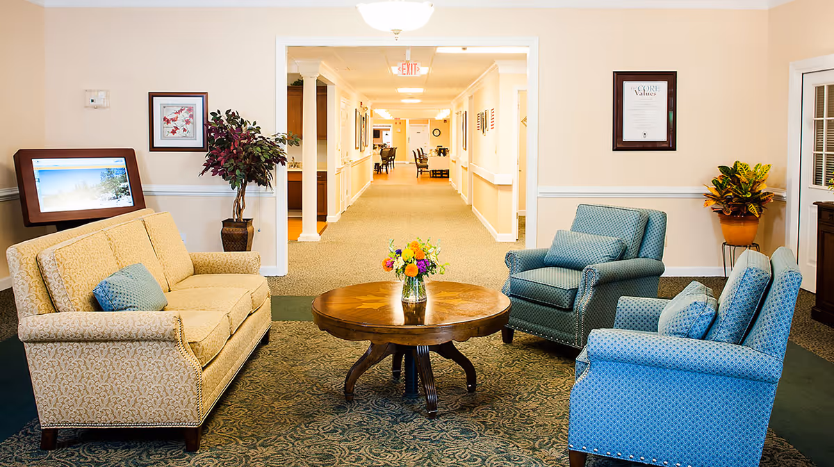Seating area in a senior living facility with a sofa, three armchairs around a round wooden coffee table and a hallway in the background.