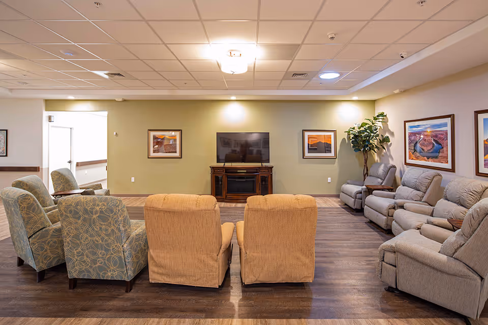 A cozy living room area in an assisted living facility with multiple upholstered armchairs and recliners arranged in a semi-circle facing a flat-screen TV mounted on a wooden stand. The walls are decorated with framed landscape photographs, and there is a tall potted plant in the corner. The room has wood flooring and a drop ceiling with recessed lighting.