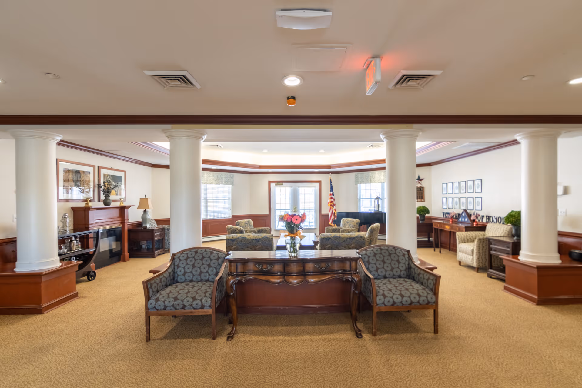 A spacious and well-lit common area in a senior living facility featuring classic wooden furniture, patterned armchairs, a wooden table with a vase of flowers, white columns, and framed artwork on the walls. There is an American flag near the windows and a piano in the background.