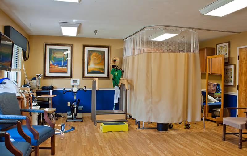 A therapy or rehabilitation room with exercise equipment including a stationary bike and parallel bars. There is a beige privacy curtain partially drawn around a section of the room. The room has wooden flooring, blue and beige walls, framed artwork, chairs, and a mirror on the right side.