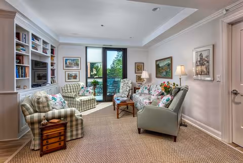 A cozy living room with patterned armchairs and a sofa arranged around a wooden coffee table. The room features built-in bookshelves filled with books and decorative items on the left wall, framed artwork on the walls, and a glass door leading to an outdoor balcony with greenery visible outside. The room is softly lit by table lamps and natural light.