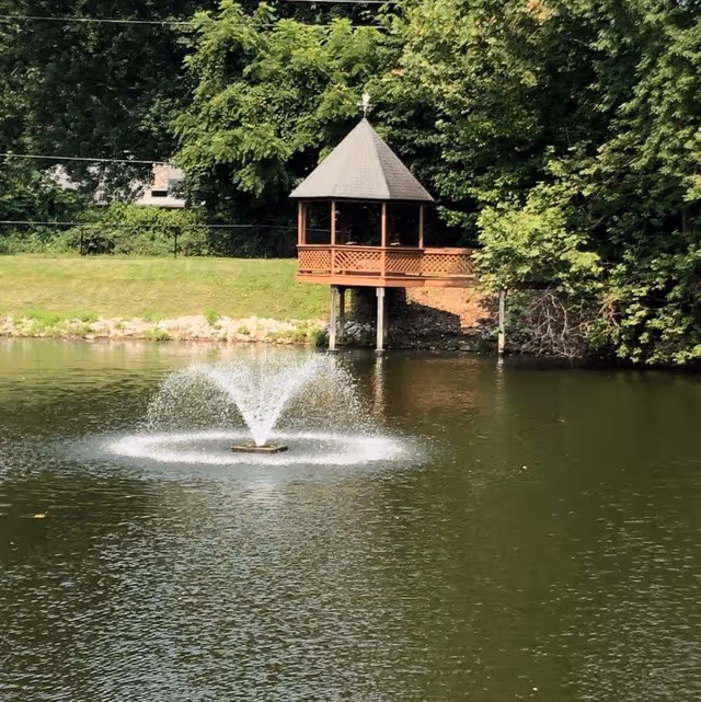 A small wooden gazebo with a pointed roof stands on stilts at the edge of a pond surrounded by green trees and grass. In the pond, a fountain sprays water upwards, creating a circular pattern on the water surface.