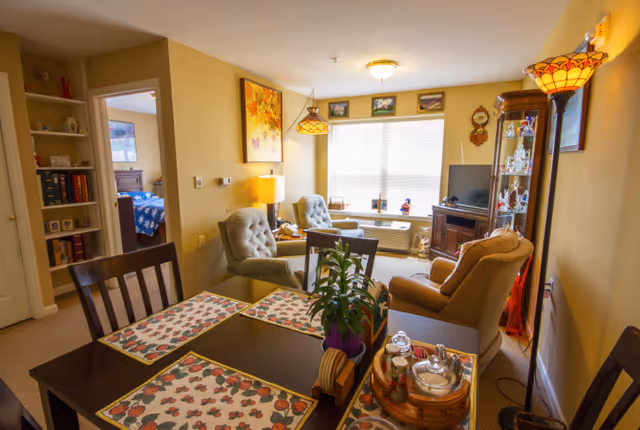 A cozy living and dining area in a senior living facility with a dark wooden dining table set with floral placemats and a potted plant centerpiece. The living room has comfortable armchairs, a TV on a wooden stand, a glass display cabinet, and a large window letting in natural light. A bedroom with a blue bedspread is visible through an open doorway.
