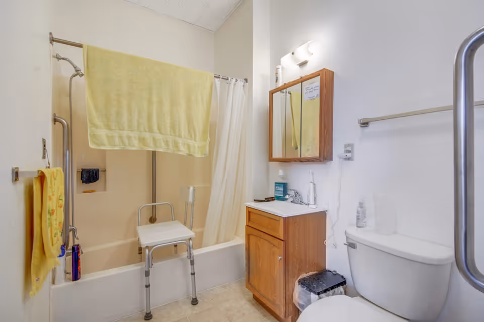 A bathroom with a bathtub equipped with a shower chair and grab bars. A yellow towel hangs over the shower curtain rod, and another yellow towel is on a wall-mounted towel rack. There is a wooden vanity with a sink, a mirror cabinet above it, and a toilet next to the vanity. Various toiletries are visible on the sink and toilet tank.