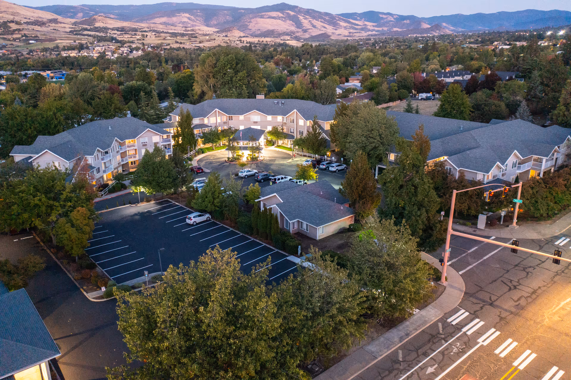 Aerial view of Maple Ridge Senior Living facility at dusk, showing multiple connected buildings surrounded by trees, a parking lot with several cars, and a nearby street intersection with traffic lights. Hills and a residential area are visible in the background.