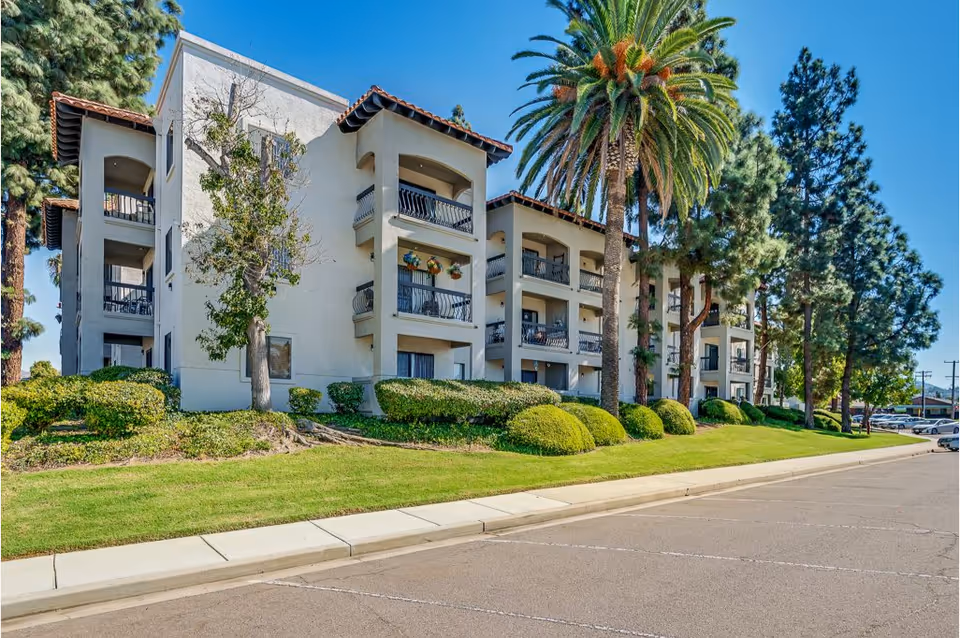Exterior view of a multi-story senior living facility building with balconies, surrounded by well-maintained landscaping including palm trees, bushes, and grass under a clear blue sky.
