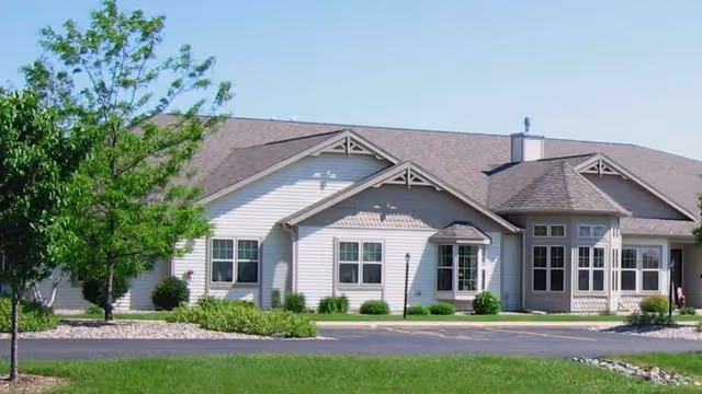 Exterior view of a single-story building with light-colored siding, multiple windows, and a shingled roof. The building is surrounded by a well-maintained lawn, small bushes, and a tree on the left side. The sky is clear and blue.