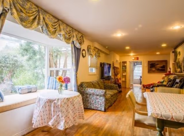 Sunlit communal living room with floral sofas, dining tables, a window seat and hardwood floors.