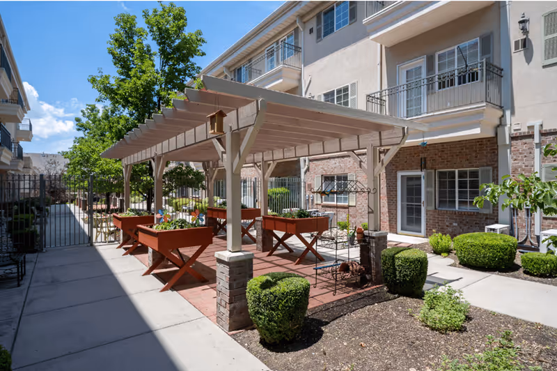Outdoor courtyard featuring a pergola with raised planter boxes and seating in front of a multi-story apartment building.