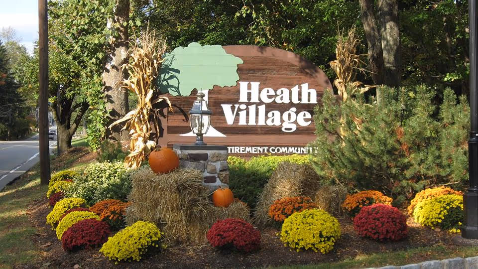 Landscaped entrance sign reading "Heath Village" with pumpkins, hay bales, corn stalks, and colorful fall mums.