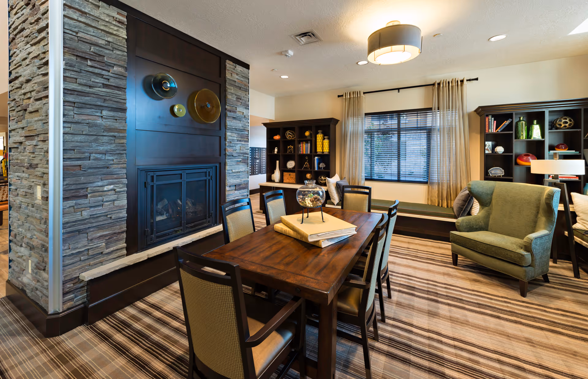 A cozy living room area featuring a wooden dining table with six chairs, a stone fireplace with decorative wall art above it, a green upholstered armchair, built-in bookshelves with various decorative items, and a window with sheer curtains allowing natural light to enter.