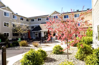 Outdoor courtyard area of a senior living facility with blooming pink trees, shrubs, and a paved walkway. The three-story beige building surrounds the courtyard, which has several tables and chairs for residents to sit and enjoy the outdoors.