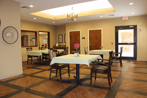 A bright dining room with several tables covered in pale yellow tablecloths, wooden chairs, potted flowers on each table, and a recessed ceiling light fixture.