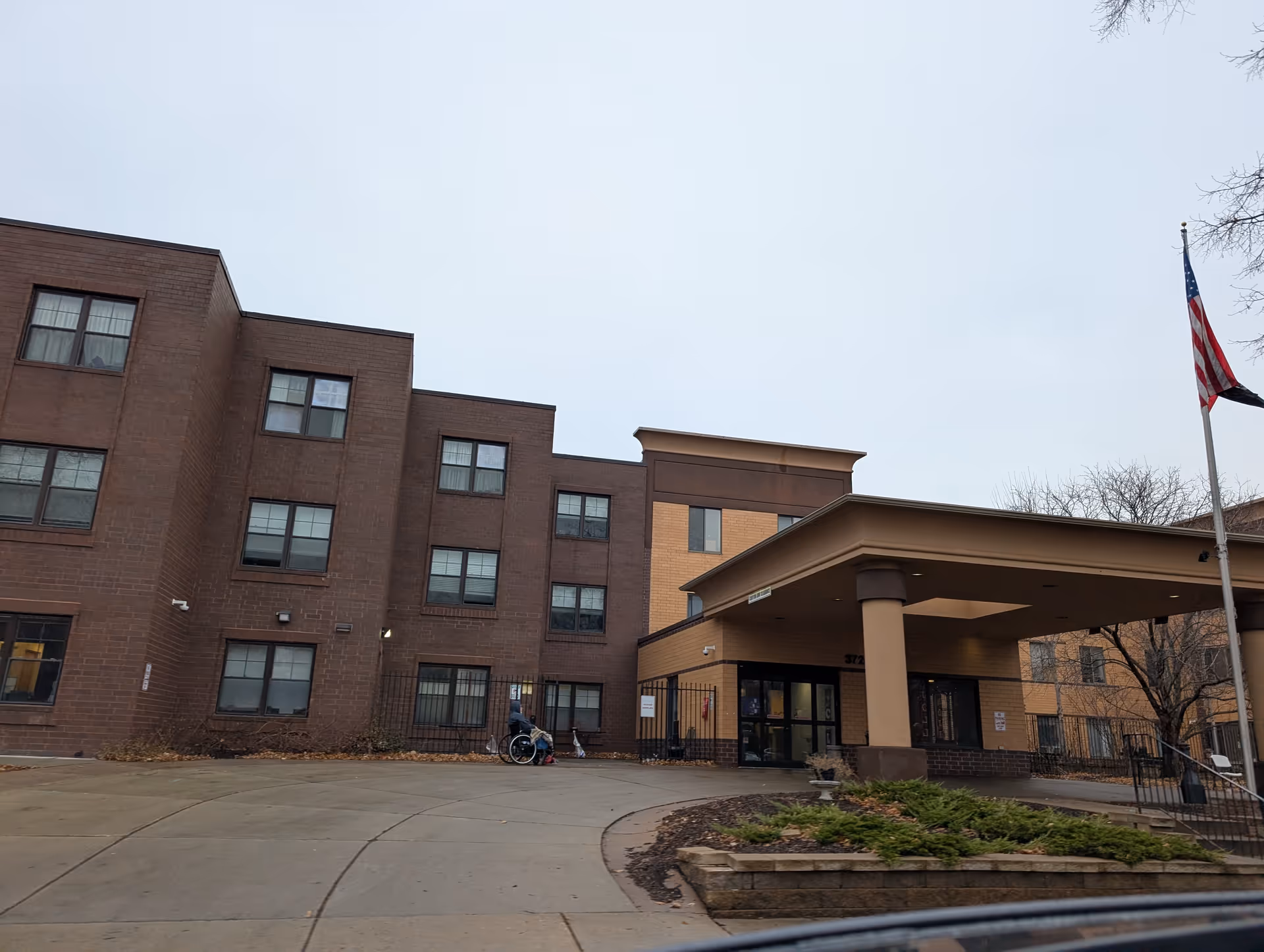Exterior view of a multi-story senior living facility building with a covered entrance, an American flag on a flagpole, and a person in a wheelchair near the entrance.