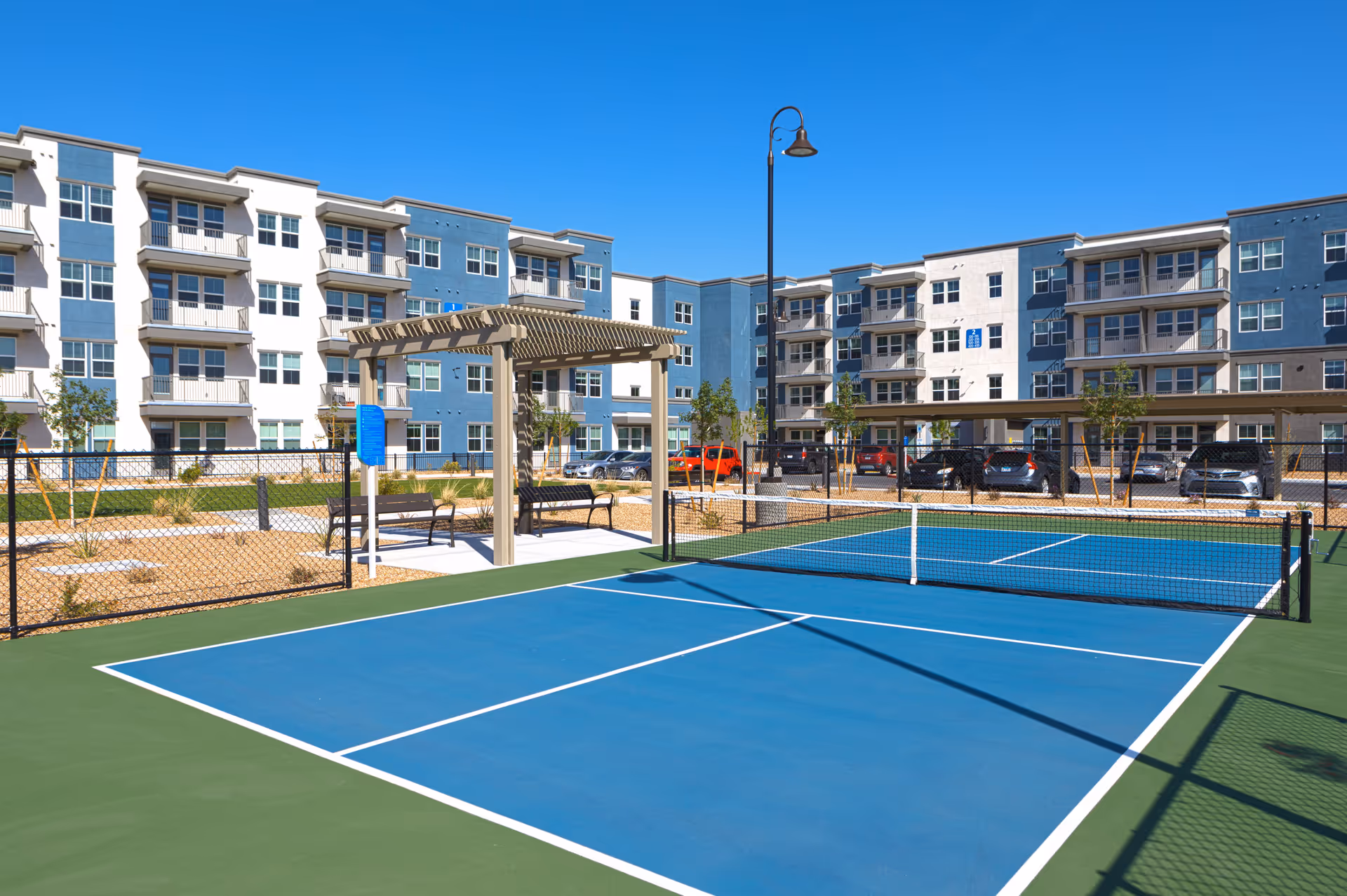 Outdoor view of a senior living facility featuring a blue and green pickleball court with a net, a pergola with benches underneath, a parking lot with cars, and a multi-story residential building with balconies in the background under a clear blue sky.