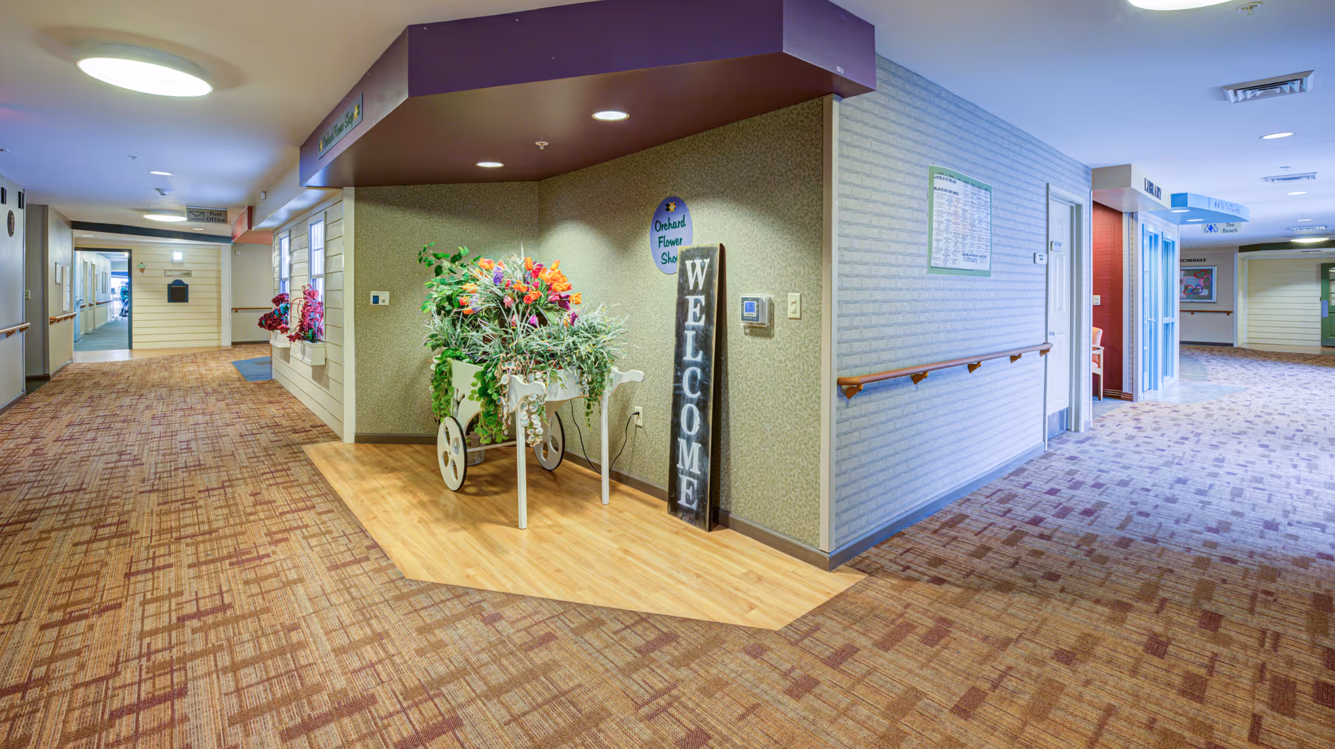 Interior hallway of Arden Courts - ProMedica Memory Care Community with patterned carpet, beige walls, handrails, and a decorative flower cart with colorful artificial flowers next to a vertical welcome sign.
