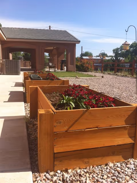 Outdoor area at GoodLife Senior Living & Memory Care featuring wooden raised garden beds with red flowers, a gravel ground, a covered patio structure, and a clear blue sky.