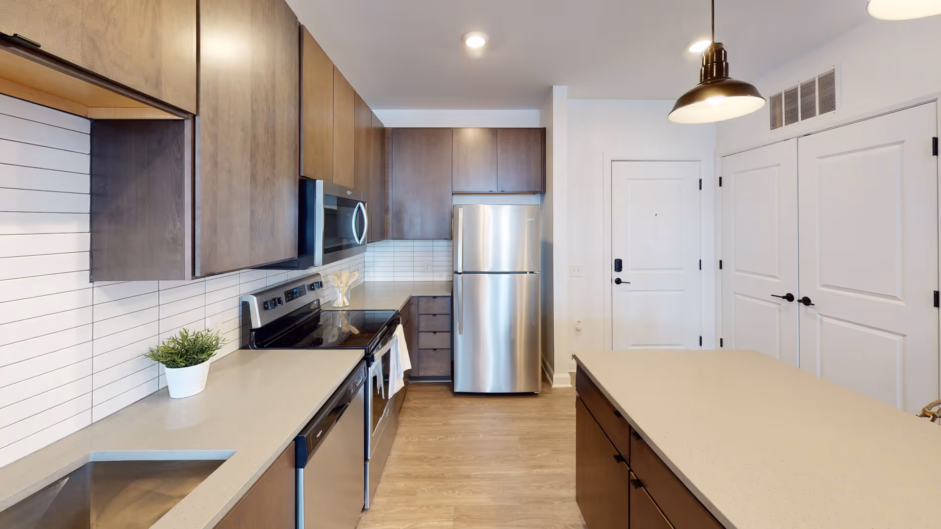 Modern kitchen with wooden cabinets, stainless steel appliances including a refrigerator, microwave, stove, and dishwasher. There is a light-colored countertop with a small potted plant, a kitchen island, and a pendant light hanging from the ceiling. The floor is wooden, and there are white walls with two closed white doors in the background.