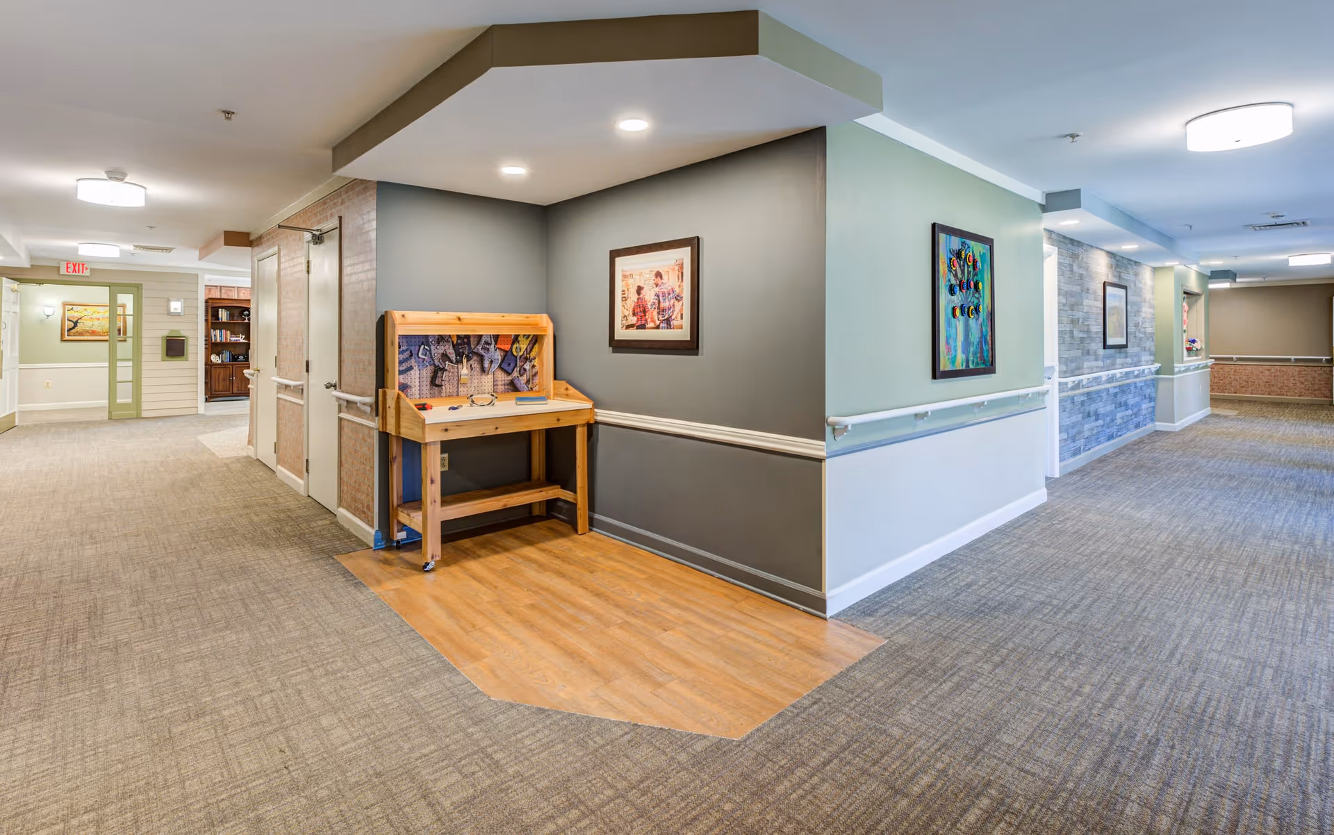 A well-lit hallway in a senior living facility with carpeted floors and light-colored walls. The hallway features handrails along the walls, framed artwork, and a small wooden activity table with tools. The ceiling has recessed lighting and a modern light fixture. Doors and an exit sign are visible at the far end of the hallway.