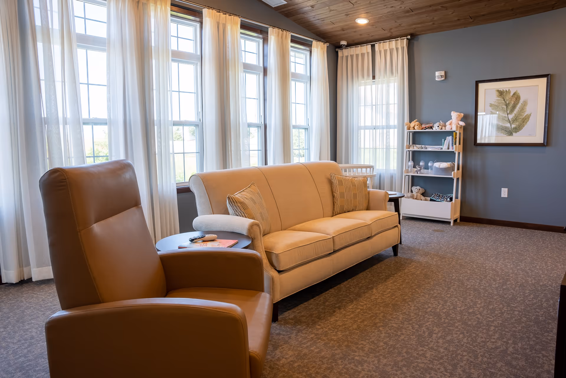 A cozy living room with a beige sofa and two striped pillows, a brown leather armchair, a small round side table with a remote and a book, large windows with sheer white curtains letting in natural light, a shelf with stuffed animals and books, and a framed botanical print on a blue-gray wall.