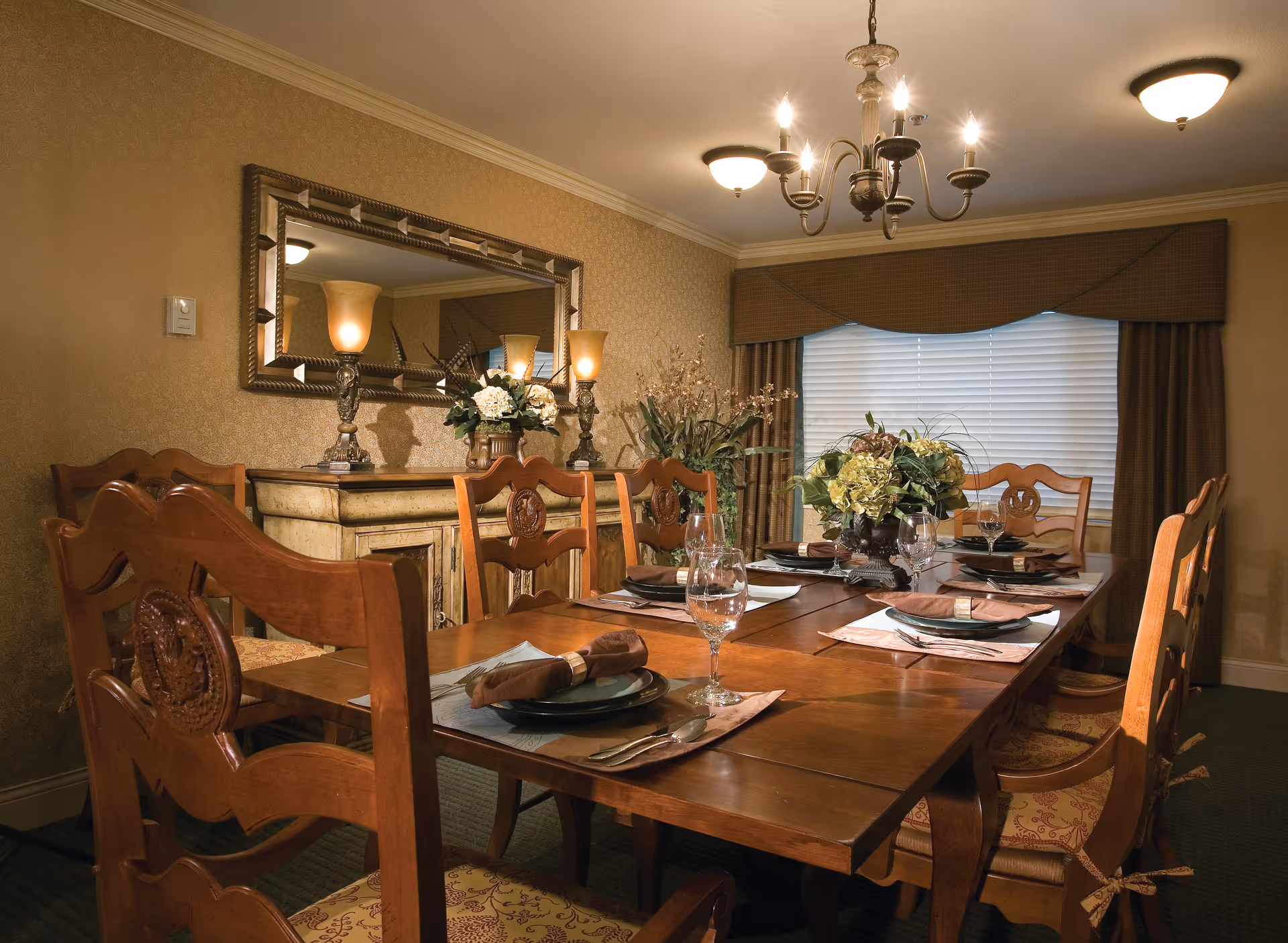 A warmly lit dining room with a wooden dining table set for six people. The table has place settings with plates, napkins, silverware, and wine glasses. There are floral centerpieces on the table. The room features a large mirror on the wall, decorative lamps, and a window with closed blinds and brown curtains. The chairs are wooden with cushioned seats.