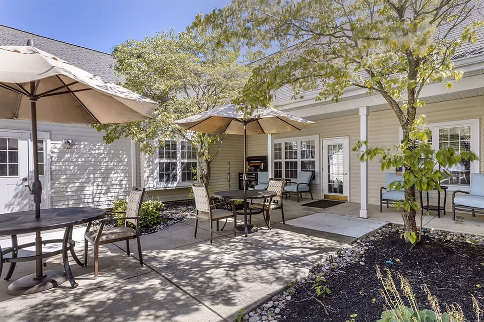 Outdoor patio area at Brookdale Dublin with round tables, chairs, and large umbrellas providing shade. The patio is surrounded by trees and plants, with a building featuring windows and doors in the background.