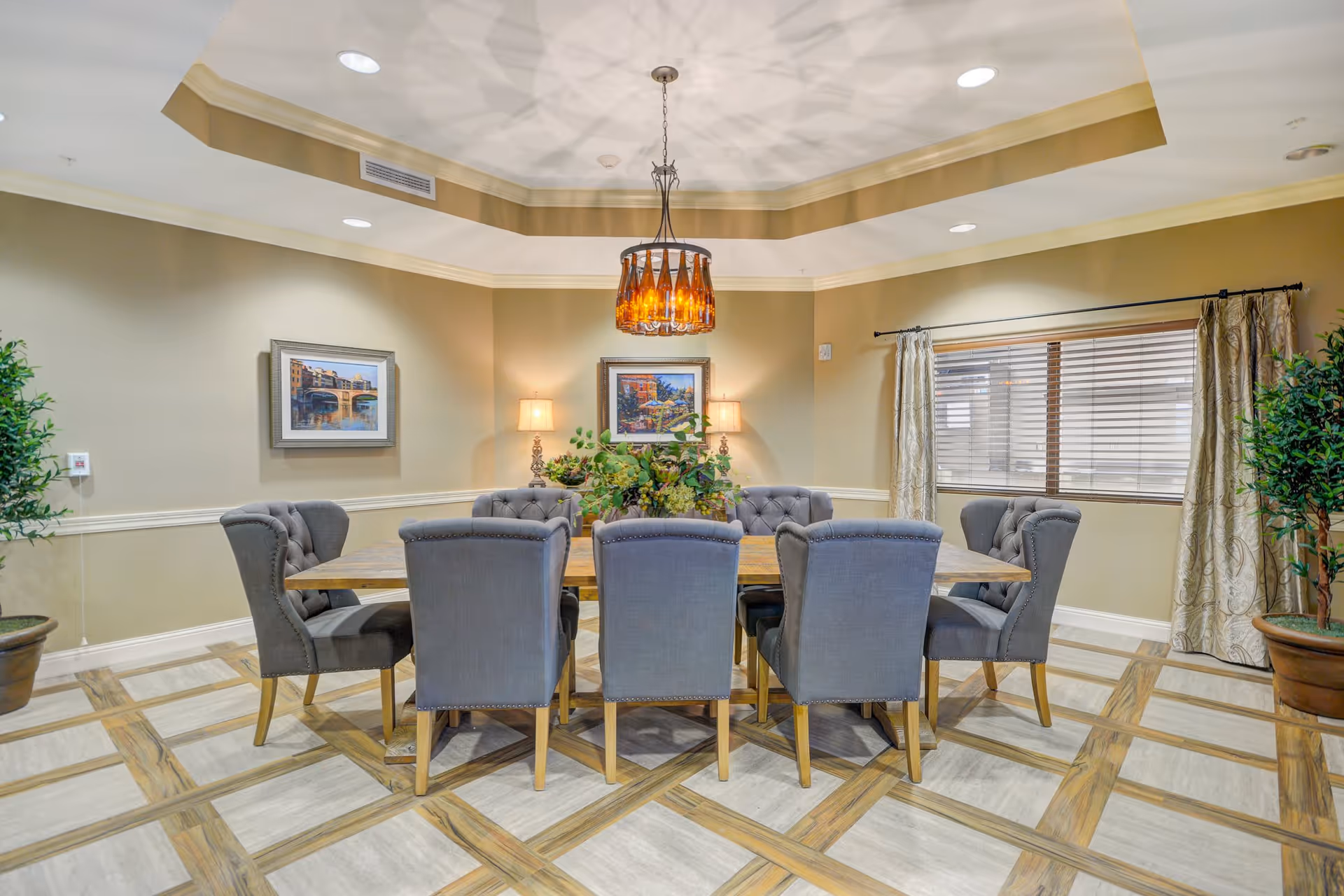 A dining room with a wooden table surrounded by eight upholstered gray chairs. The room features a decorative ceiling light fixture, two framed paintings on the walls, two table lamps, large window with curtains, and potted plants in the corners. The floor has a patterned tile design.