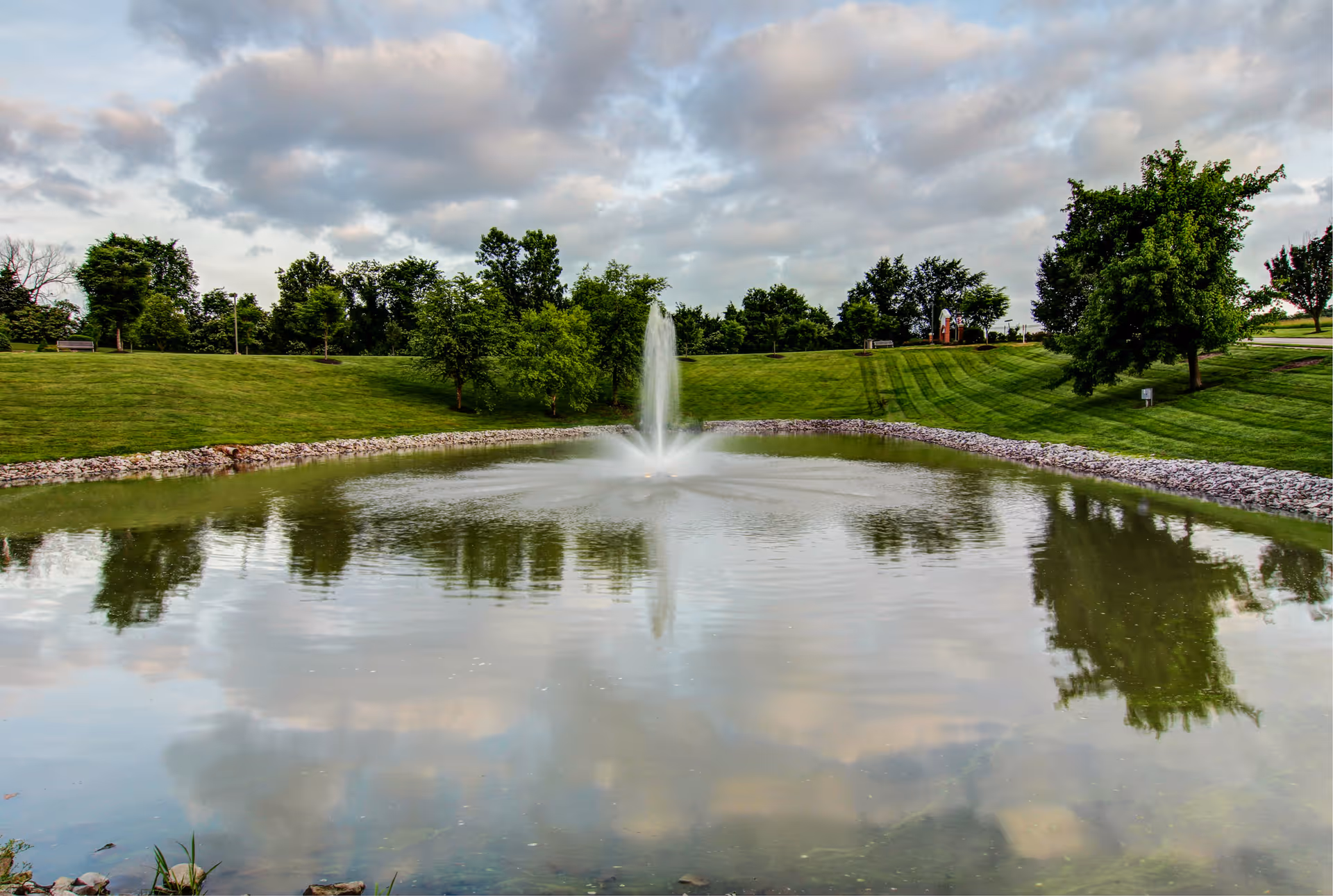 A serene outdoor scene featuring a pond with a central water fountain surrounded by green grass and trees under a partly cloudy sky.