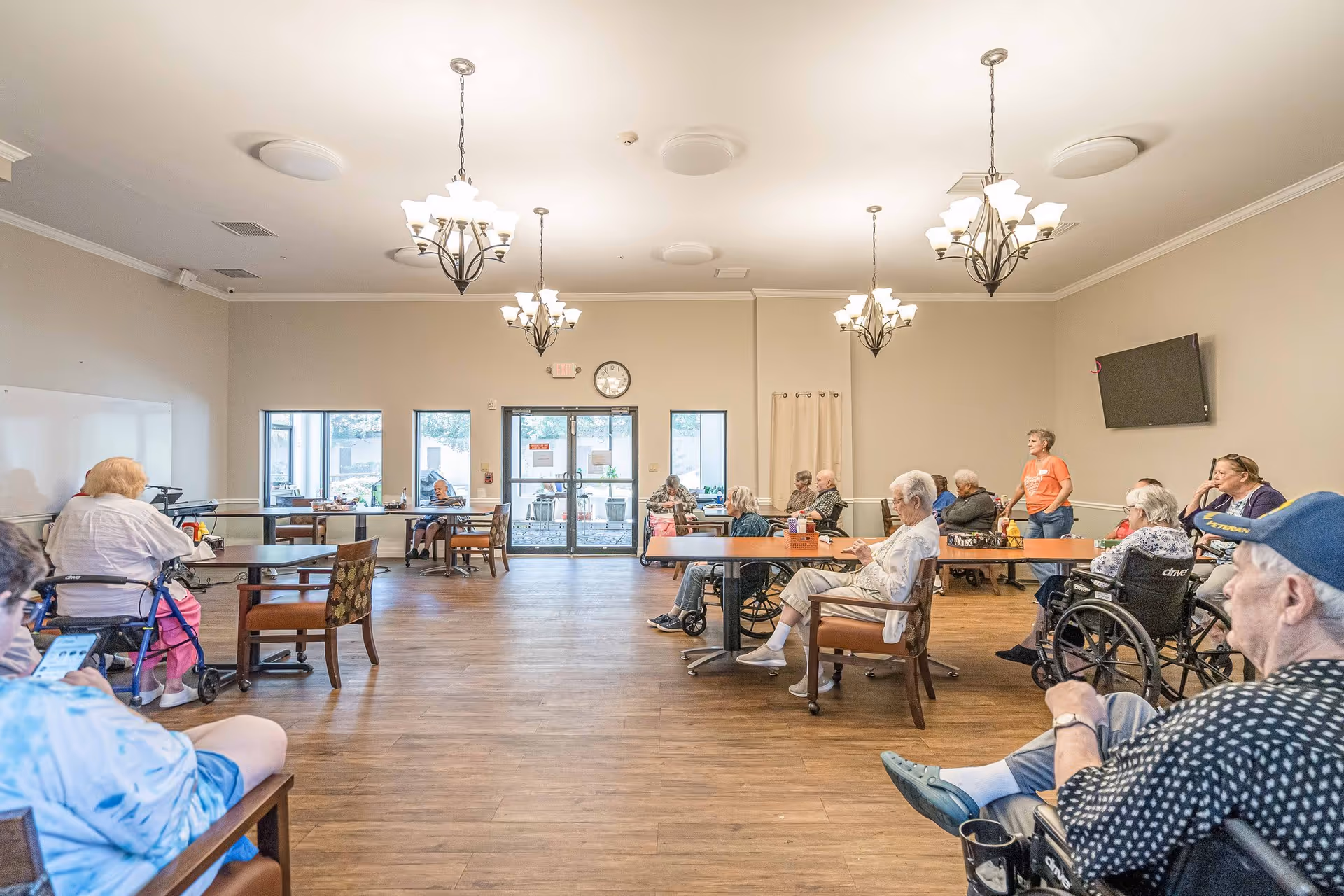 A spacious room with wooden flooring and beige walls, featuring several elderly individuals seated around tables and in wheelchairs. The room has multiple chandeliers hanging from the ceiling, a wall-mounted TV, and large windows and doors at the far end letting in natural light. A staff member in an orange shirt stands near the back of the room.