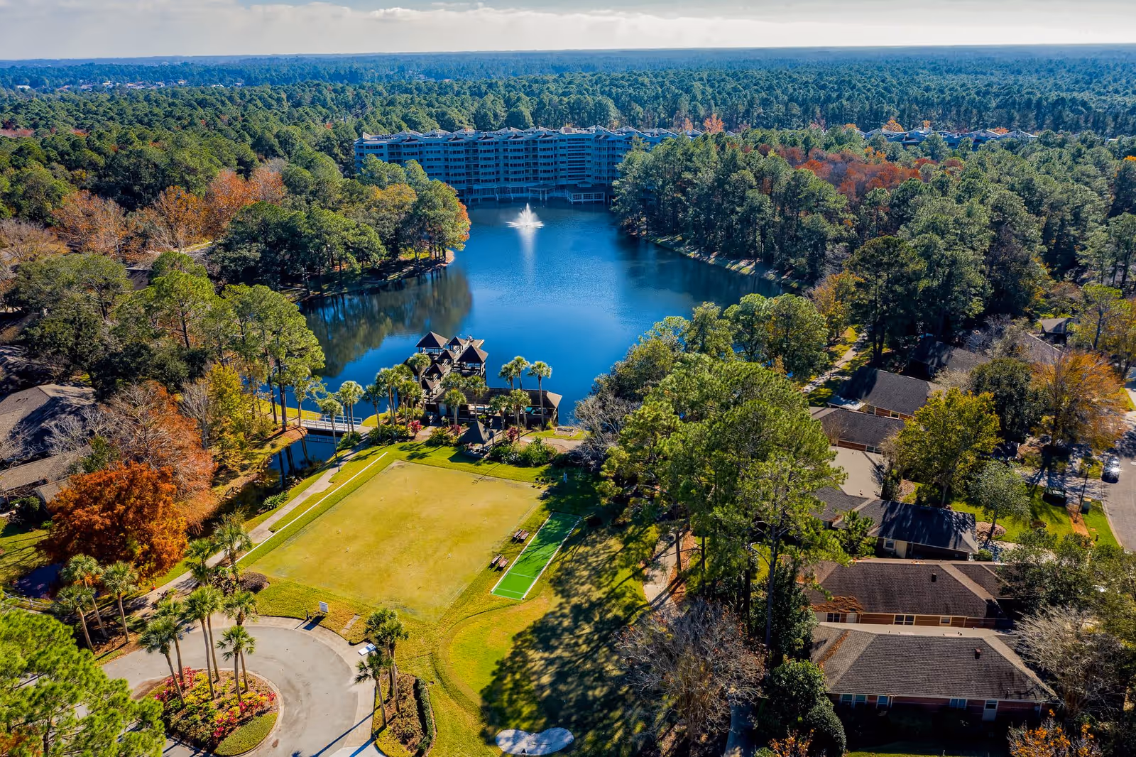 Aerial view of Cypress Village showing a large pond with a water fountain in the center, surrounded by trees and greenery. There are multiple buildings around the pond, including a large multi-story building in the background and smaller residential-style buildings in the foreground. A well-maintained lawn area with pathways and palm trees is visible near the pond.