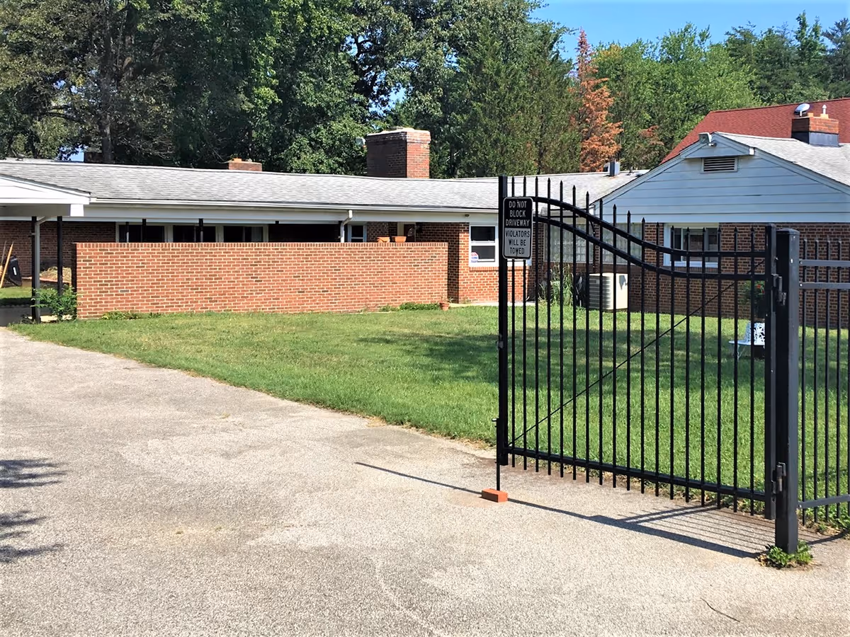 Exterior view of a single-story brick building with a gray roof, surrounded by green grass and trees. A black metal gate is partially open in the foreground with a sign that reads 'DO NOT BLOCK DRIVEWAY VIOLATORS WILL BE TOWED.'