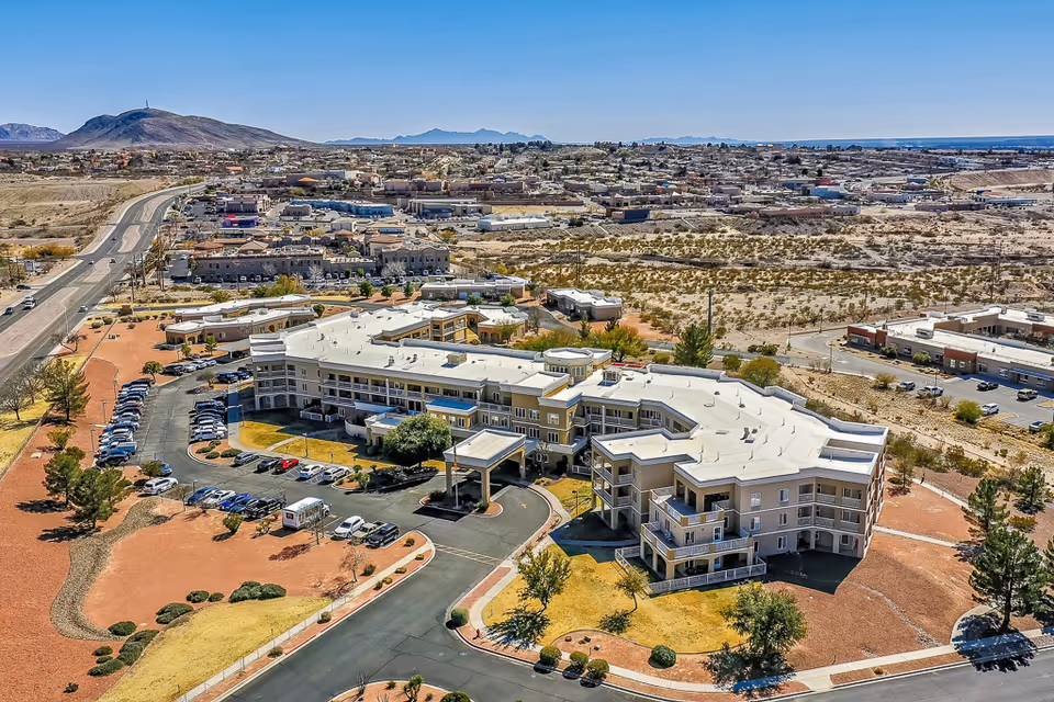 Aerial view of Solstice Senior Living at Las Cruces, a large multi-story senior living facility surrounded by parking lots, roads, and desert landscape with mountains in the background under a clear blue sky.