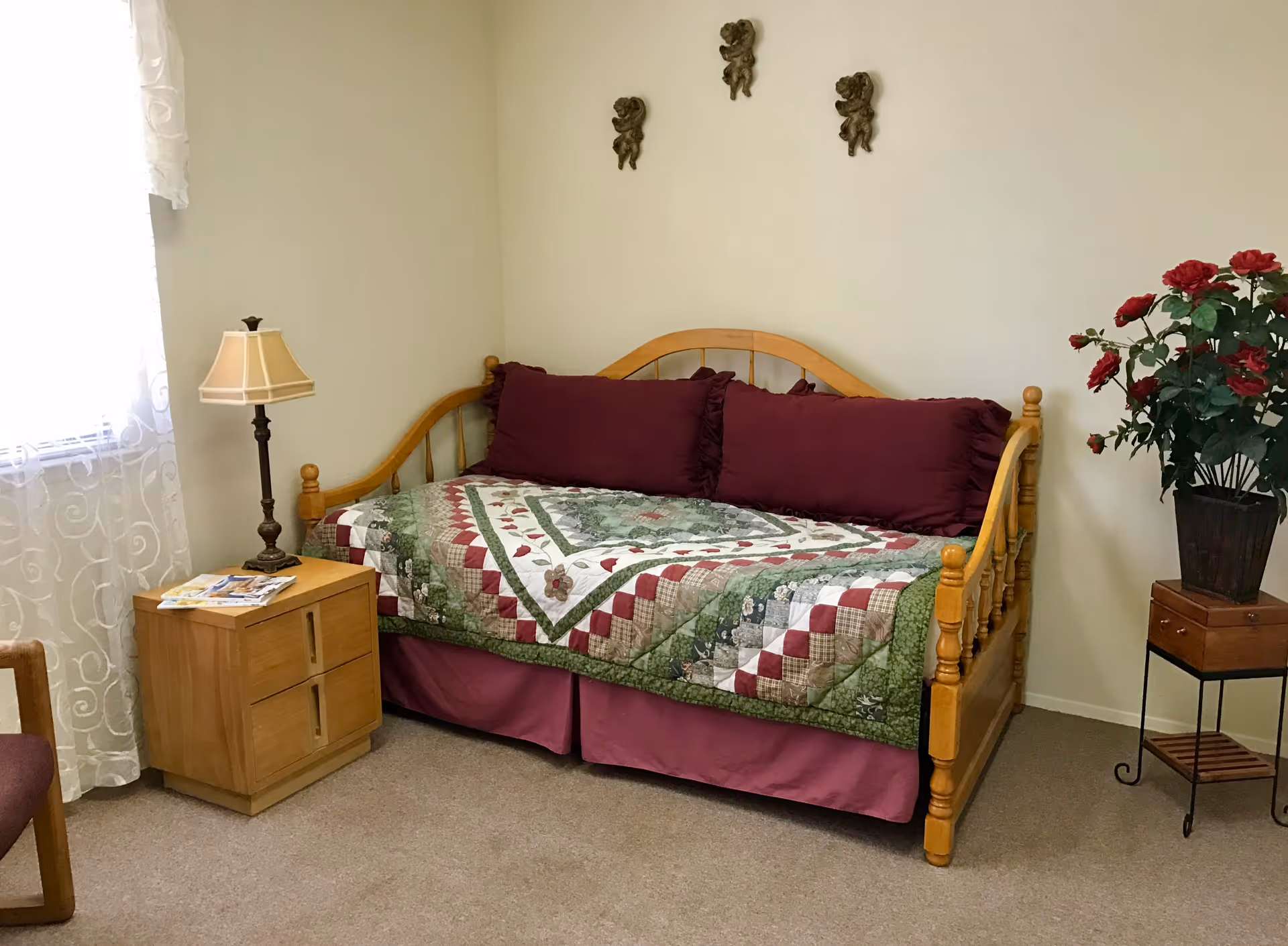 A cozy room with a wooden daybed covered with a green, red, and white patchwork quilt and three large burgundy pillows. To the left of the bed is a wooden nightstand with a table lamp and some magazines. A window with sheer white curtains is partially visible. On the right side, there is a tall plant with red flowers in a black pot on a small wooden stand. The walls are light-colored with three small decorative cherub wall hangings above the bed.