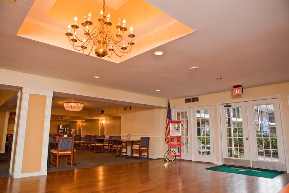 Interior view of a senior living facility dining area with wooden floors, multiple tables and chairs, a chandelier hanging from a recessed ceiling, and glass doors leading outside. An American flag and a red popcorn machine are positioned near the doors.