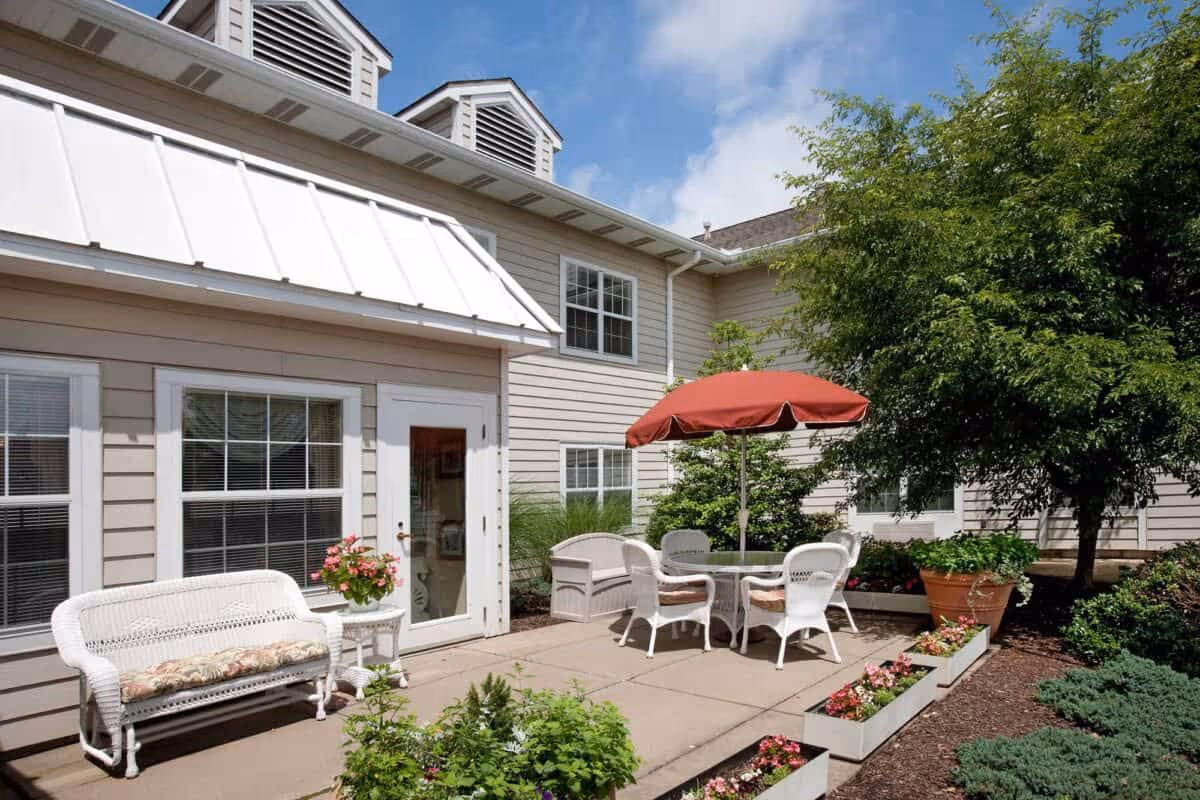 Outdoor courtyard patio with white wicker seating, a table under a red umbrella, potted plants and flower boxes beside a beige two-story building.