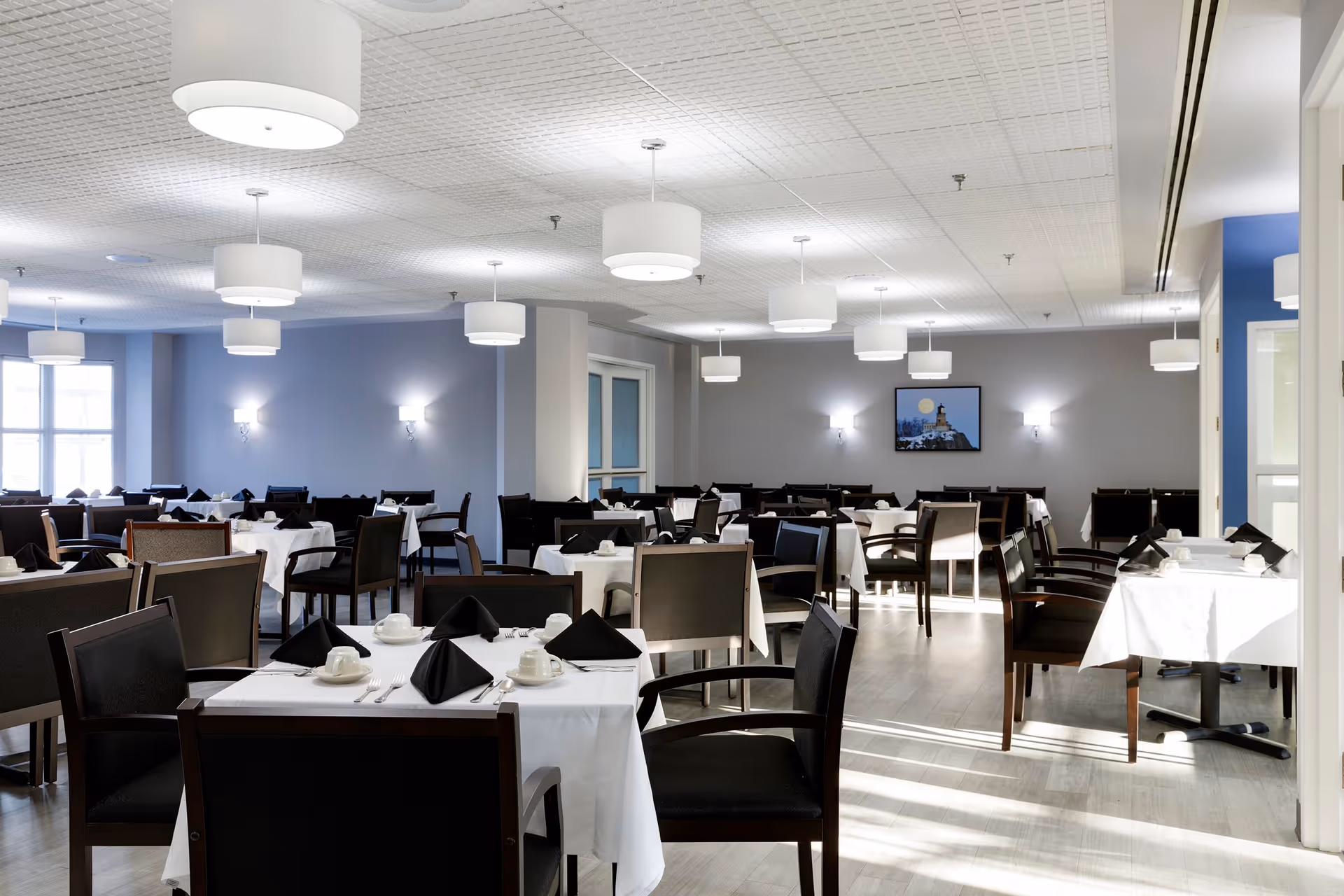A spacious dining room with multiple tables covered in white tablecloths, each set with black napkins, white cups, and silverware. The room has modern lighting fixtures hanging from the ceiling and wall sconces providing additional light. There is a framed picture on the far wall and large windows letting in natural light.
