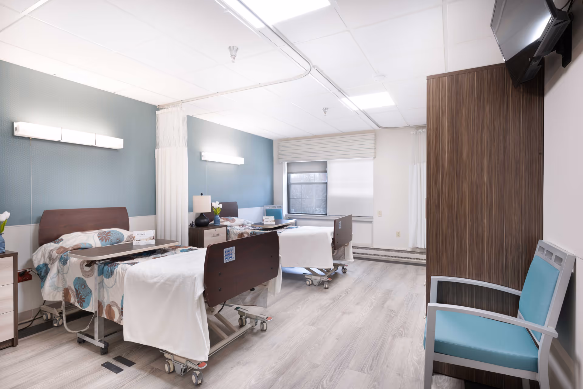 A bright and clean nursing facility room with two hospital beds, each with a floral patterned blanket and a white towel. There are bedside tables with lamps and flowers, a window with blinds, a mounted TV on the wall, and a blue cushioned chair near a wooden partition.